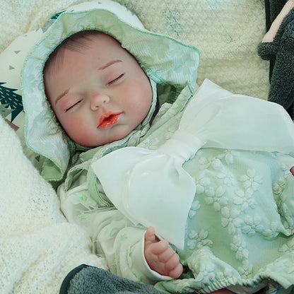 A reborn baby doll dressed in a green outfit with a hood, peacefully sleeping on a light blanket.