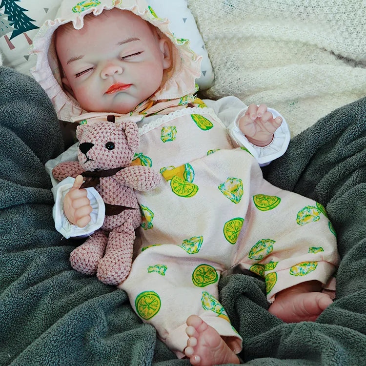 A reborn baby doll dressed in a light-colored outfit with lemon prints, peacefully sleeping on a dark blanket, holding a pink teddy bear.