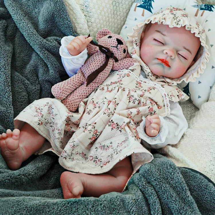 A sleeping reborn baby doll dressed in a floral bonnet and matching outfit, lying on a soft blanket and holding a knitted teddy bear.