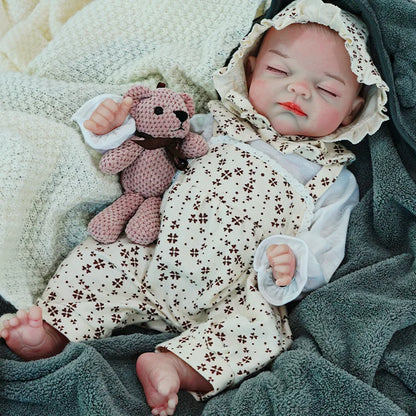  A sleeping reborn baby doll lying on a blanket, dressed in a floral onesie with a matching bonnet, holding a knitted teddy bear.