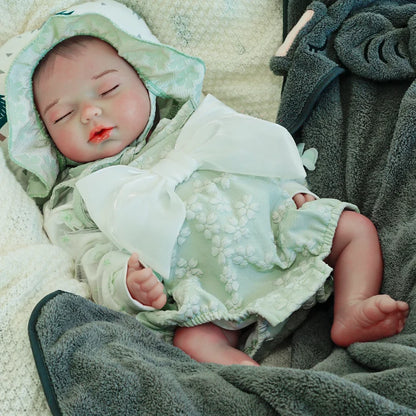 A reborn baby doll in a green hooded outfit, lying on a dark blanket with eyes closed