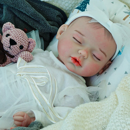 A reborn baby doll dressed in a white outfit, lying on a dark blanket, peacefully sleeping with eyes closed.