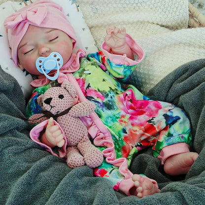 A reborn baby doll dressed in a colorful outfit with a pink headband, lying on a dark blanket with eyes closed, holding a pink teddy bear.