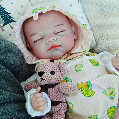 A reborn baby doll in a lemon-printed outfit, lying on a dark blanket, peacefully sleeping with eyes closed, holding a pink teddy bear.