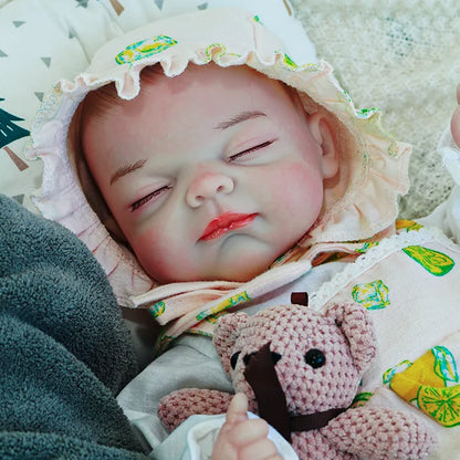 A close-up of a reborn baby doll in a lemon-printed outfit, sleeping peacefully while holding a pink teddy bear.