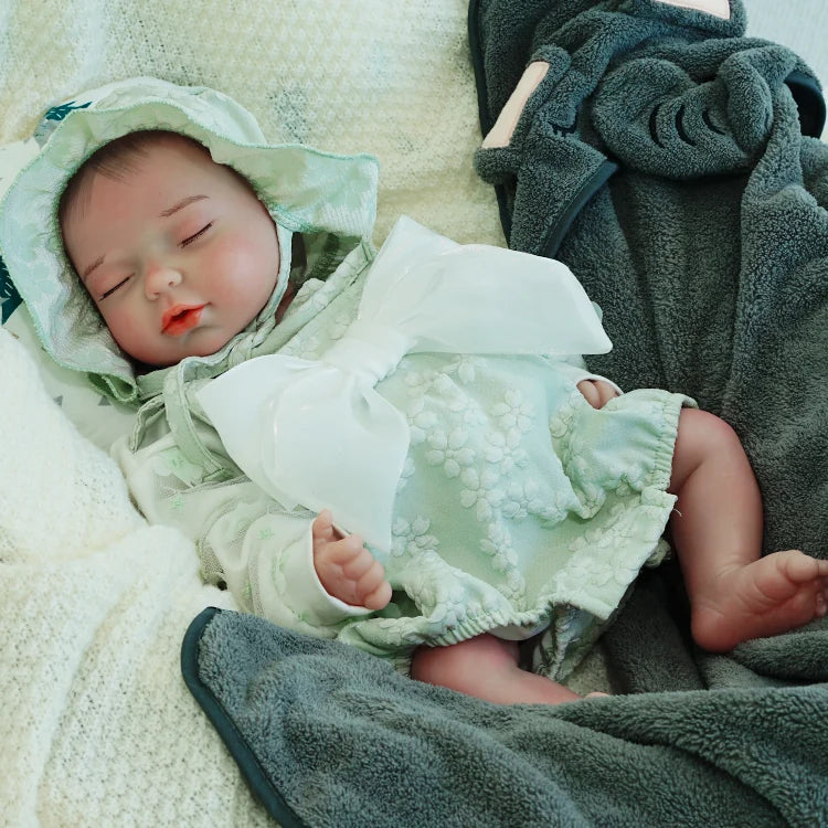  A reborn baby doll resting on a dark blanket, dressed in a green outfit with a hood.