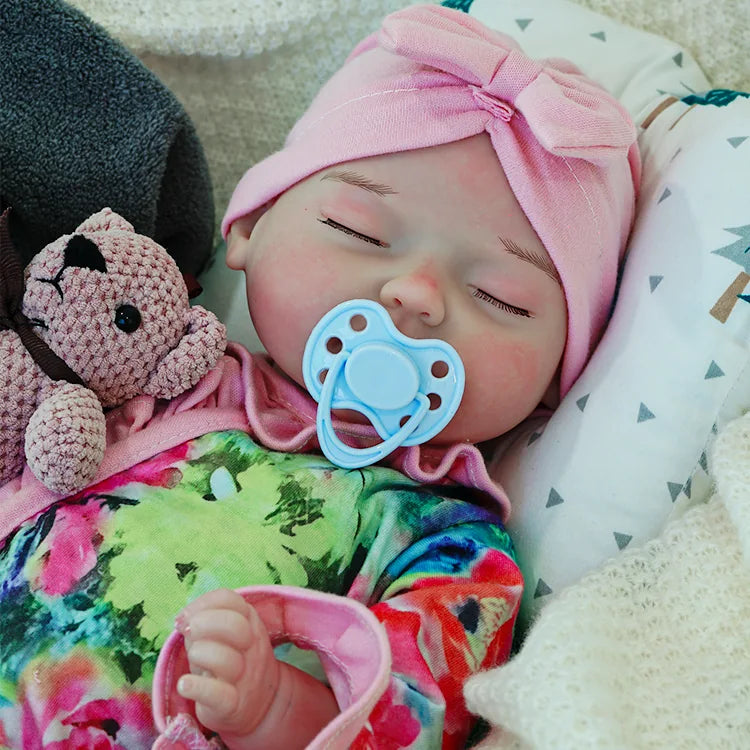 A reborn baby doll in a colorful outfit with a pink headband, peacefully sleeping while holding a pink teddy bear.