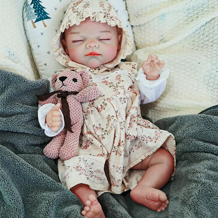 The reborn baby doll resting with eyes closed, dressed in a floral outfit and bonnet, clutching a knitted teddy bear.