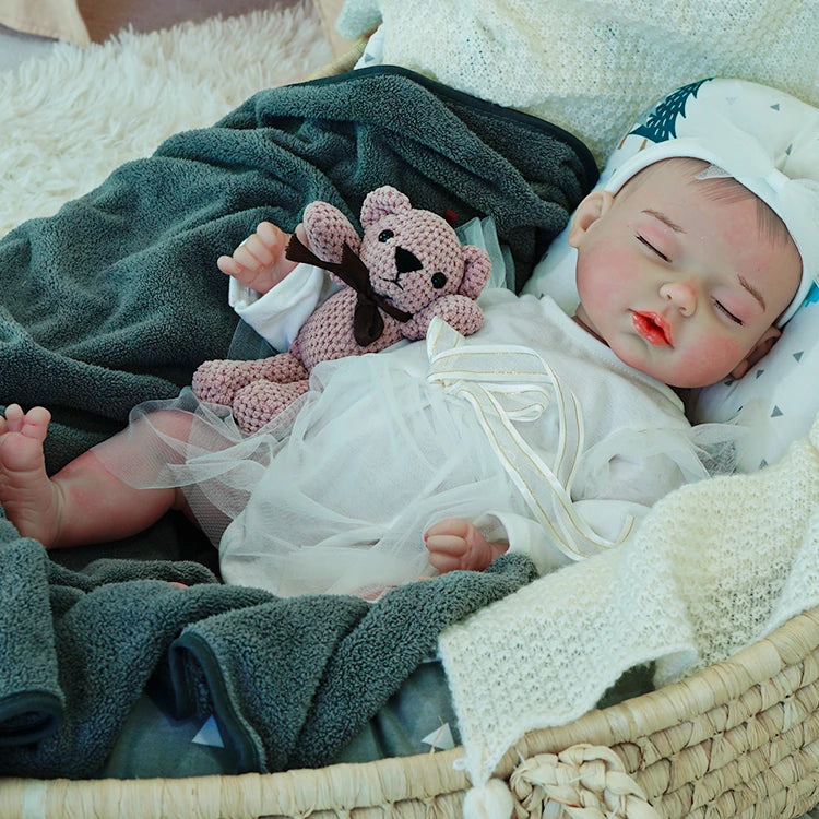 A reborn baby doll in a white outfit, resting on a dark blanket, holding a pink teddy bear.