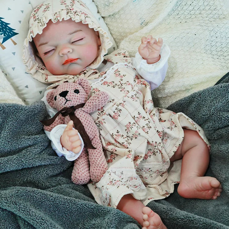 The reborn baby doll lying peacefully on a blanket, wearing a floral bonnet and dress, with a soft teddy bear in hand.