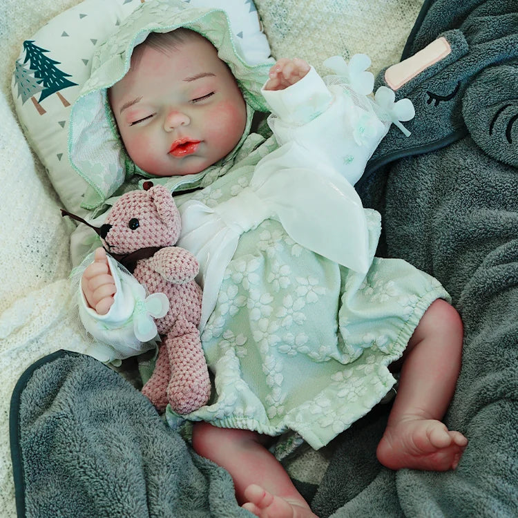 A reborn baby doll dressed in a green hooded outfit, holding a pink teddy bear, lying on a dark blanket.