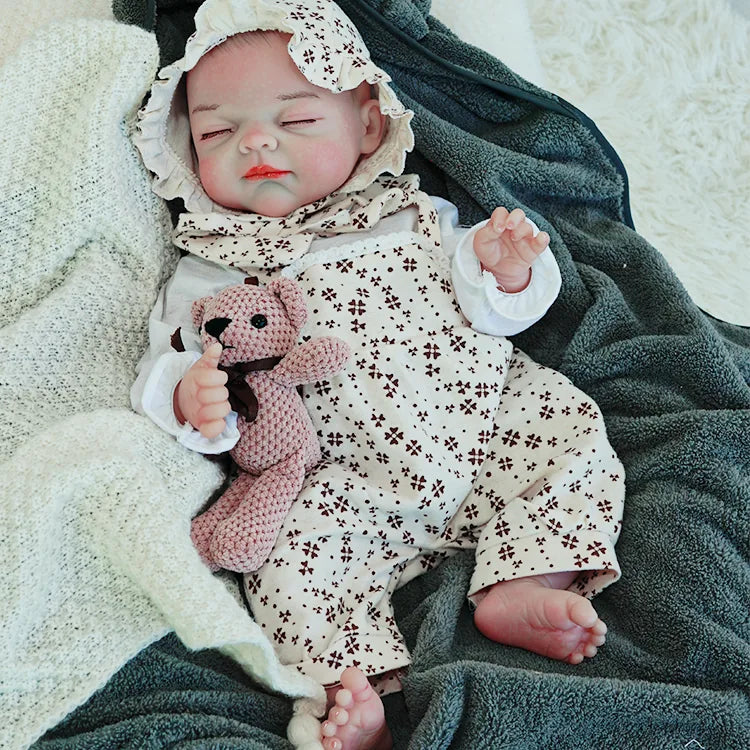 The reborn baby doll lying on a soft blanket, dressed in a floral outfit with a matching bonnet, clutching a small teddy bear.