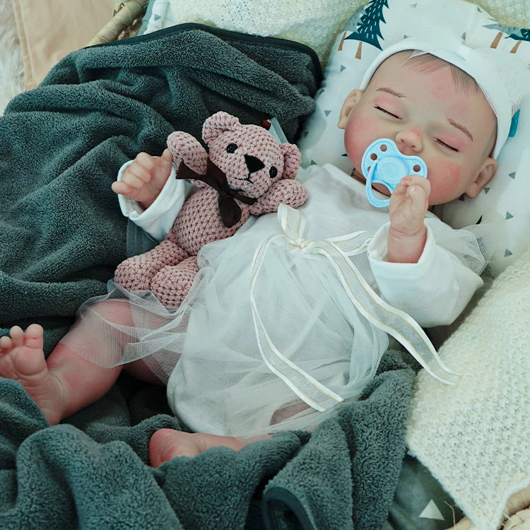 A reborn baby doll with a pacifier, dressed in white, lying on a dark blanket with a pink teddy bear.