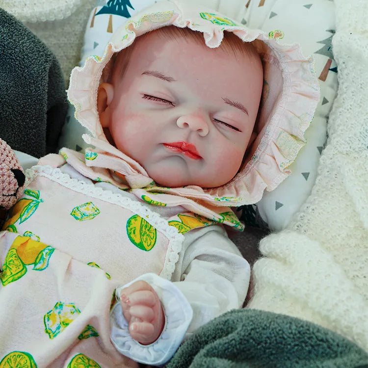 A reborn baby doll dressed in a lemon-printed outfit, lying on a dark blanket, holding a pink teddy bear.