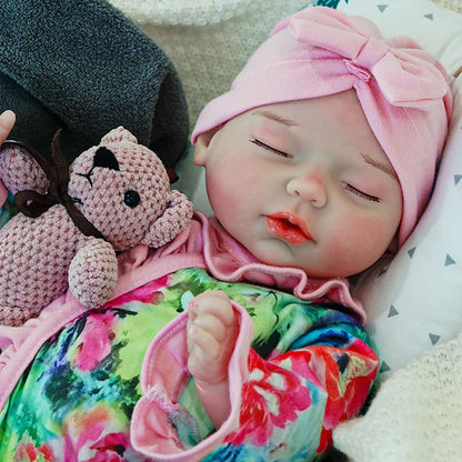 A reborn baby doll with a pacifier, dressed in a colorful outfit with a pink headband, lying on a dark blanket, holding a pink teddy bear.