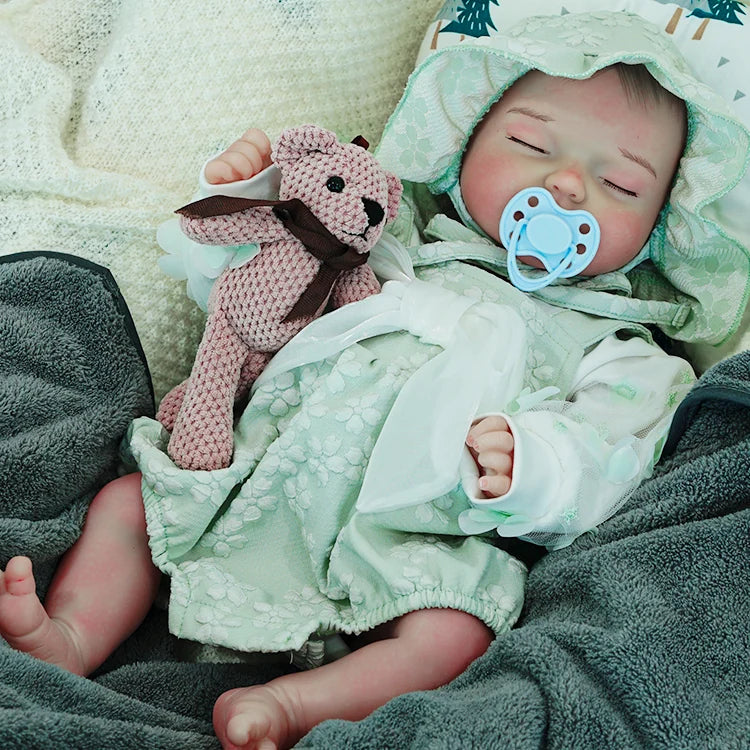 A reborn baby doll with a pacifier, dressed in a green hooded outfit, holding a pink teddy bear.