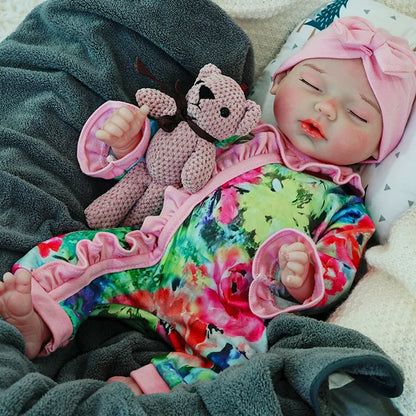 A reborn baby doll dressed in a colorful outfit with a pink headband, peacefully sleeping on a dark blanket, holding a pink teddy bear.