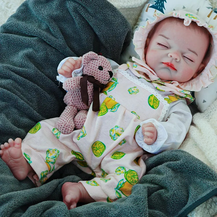 A reborn baby doll lying on a dark blanket, dressed in a lemon-printed outfit, holding a pink teddy bear.