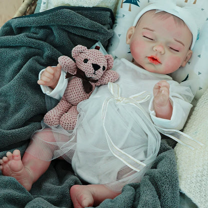 A reborn baby doll lying on a dark blanket, dressed in a white outfit, holding a pink teddy bear.