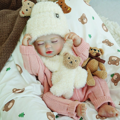 Sleeping reborn baby doll with a sheep hat, holding two toy bears, lying on a bed with bear-patterned sheets.