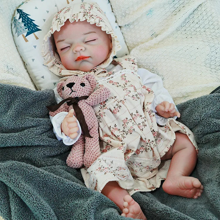  The reborn baby doll lying comfortably, dressed in a floral bonnet and outfit, with a knitted teddy bear beside her.