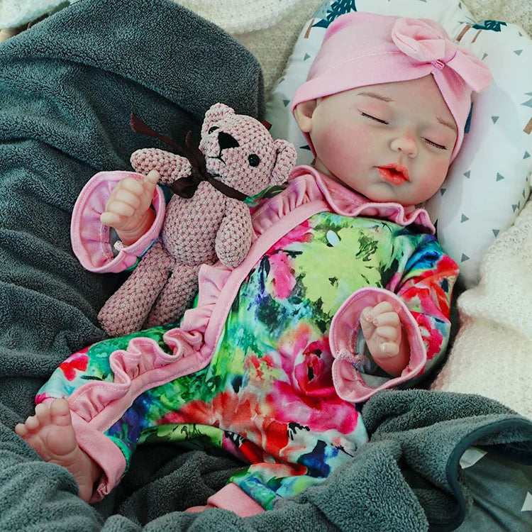 A reborn baby doll with a pacifier, dressed in a colorful outfit and a pink headband, lying on a dark blanket, holding a pink teddy bear.