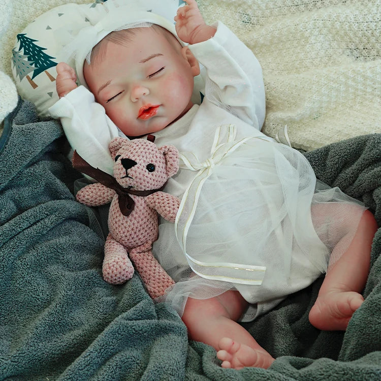 A reborn baby doll dressed in white, lying on a dark blanket with a pink teddy bear, arms raised.