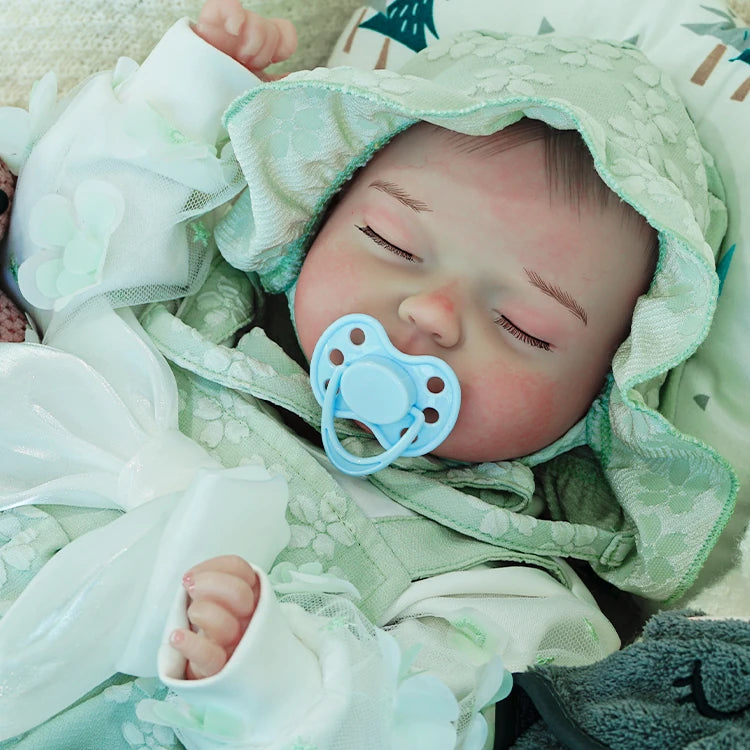 A reborn baby doll with a pacifier, dressed in a green hooded outfit, lying on a light blanket.