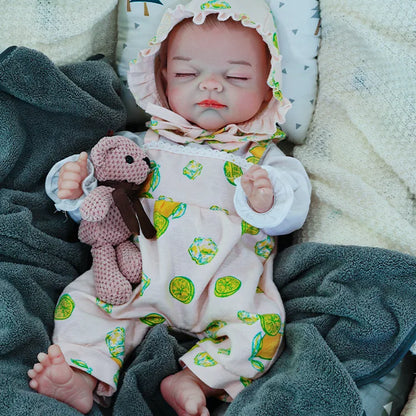 A reborn baby doll dressed in a lemon-printed outfit, lying on a dark blanket with eyes closed, holding a pink teddy bear.