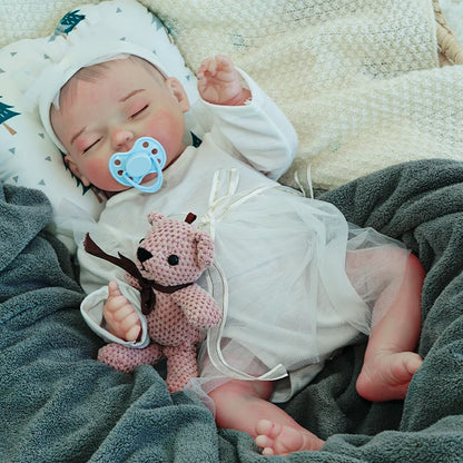 A reborn baby doll with a pacifier, dressed in white, lying on a dark blanket, holding a pink teddy bear.