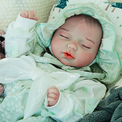 A reborn baby doll sleeping peacefully, dressed in a green hooded outfit, resting on a light blanket.