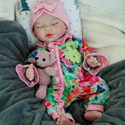 A reborn baby doll in a colorful outfit with a pink headband, peacefully sleeping on a pillow with a pacifier in its mouth, holding a pink teddy bear.