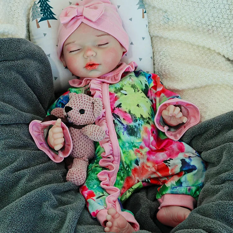 A reborn baby doll in a colorful outfit with a pink headband, peacefully sleeping on a pillow with a pacifier in its mouth, holding a pink teddy bear.