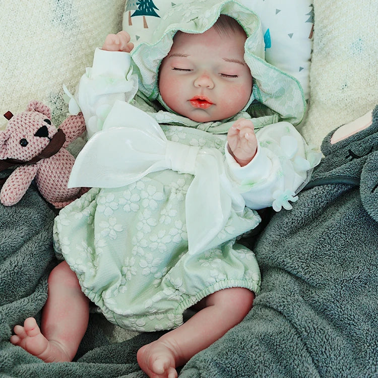 A reborn baby doll lying on a dark blanket, dressed in a green hooded outfit, holding a pink teddy bear.