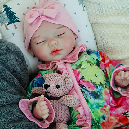 A close-up of a reborn baby doll dressed in a colorful outfit with a pink headband, sleeping with a pink teddy bear.