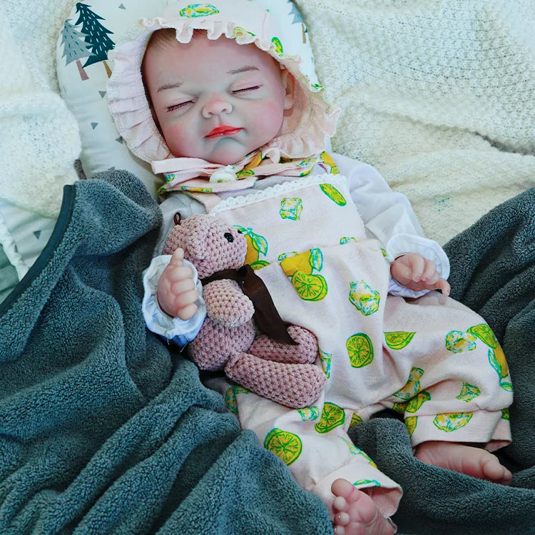 A reborn baby doll in a lemon-printed outfit, peacefully sleeping on a dark blanket while holding a pink teddy bear.