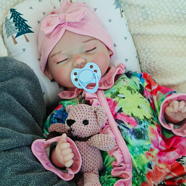 A reborn baby doll in a colorful outfit with a pink headband, lying on a dark blanket, holding a pink teddy bear.