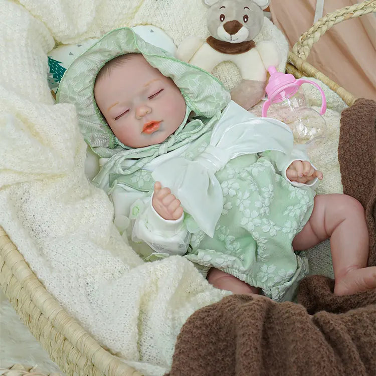 A peaceful reborn baby doll with a pink pacifier, clothed in a pale green floral hooded outfit, raising its tiny hand while resting in a wicker basket lined with a cozy white blanket, accompanied by a plush grey teddy bear and a pink feeding bottle.