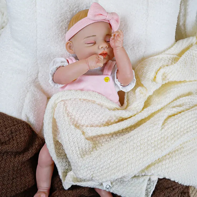 Realistic reborn baby girl doll dressed in a pink pinafore and white blouse, complete with a pacifier on the side, taking a nap on a cozy blanket.
