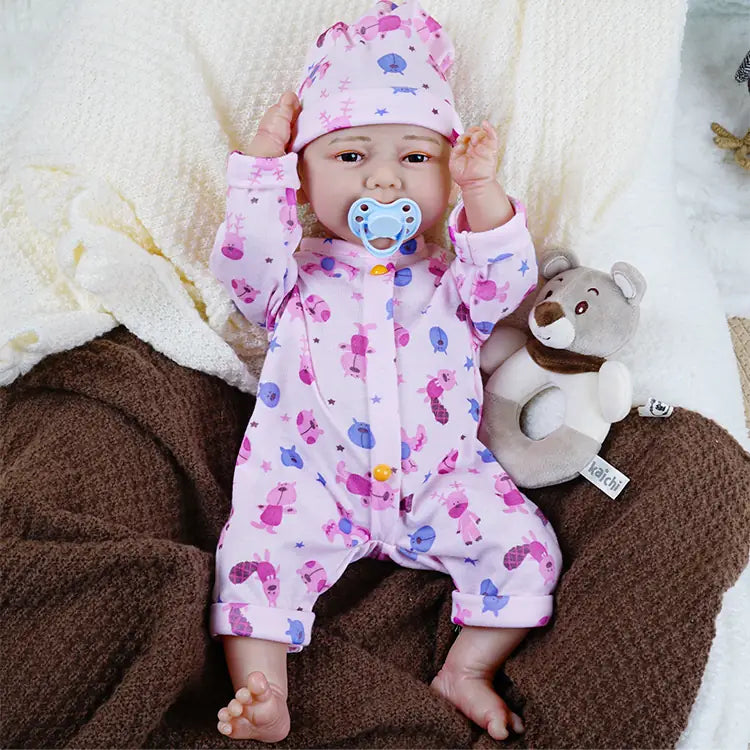 Smiling reborn baby doll in pink sea-themed pajamas seated with plush bear toys, on a cream blanket beside a brown blanket