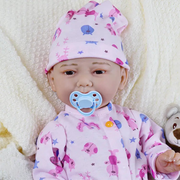 Happy reborn baby doll in pink pajamas with sea creatures, holding a dummy and a bottle, sitting on a white fluffy blanket.