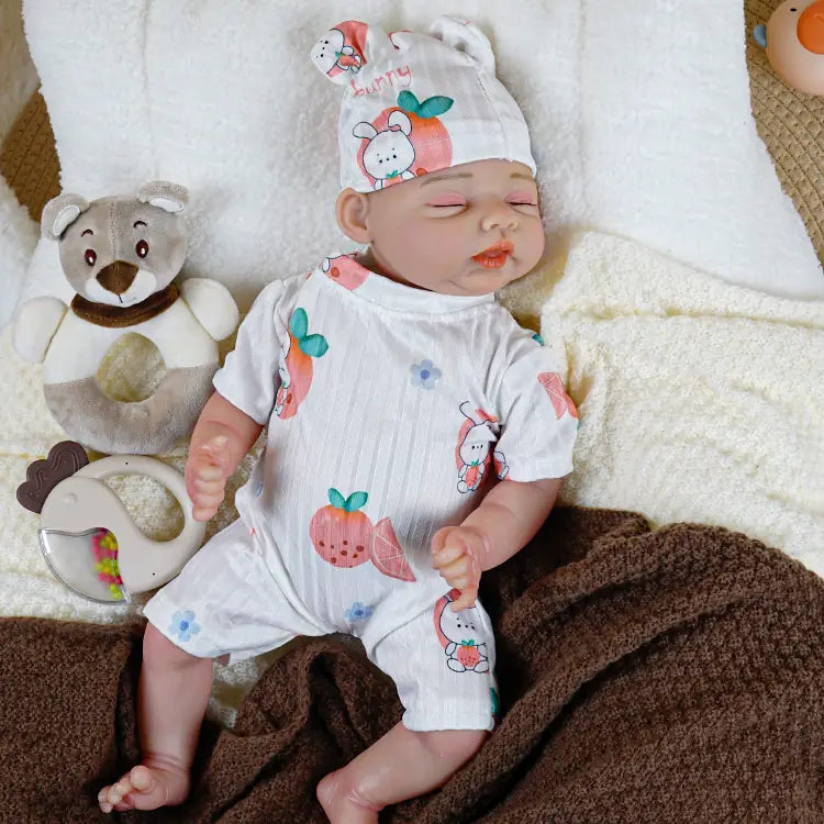 A sleeping reborn baby doll with a fruit-patterned white romper and hat, surrounded by a cozy blanket and cuddly plush toys, capturing a serene naptime scene.