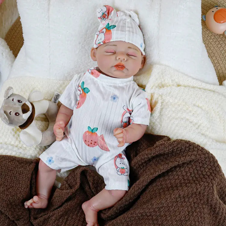 Lifelike reborn baby doll dressed in a white, fruit-print romper and a matching "bunny" hat, lying next to a plush koala toy, with a teething ring nearby
