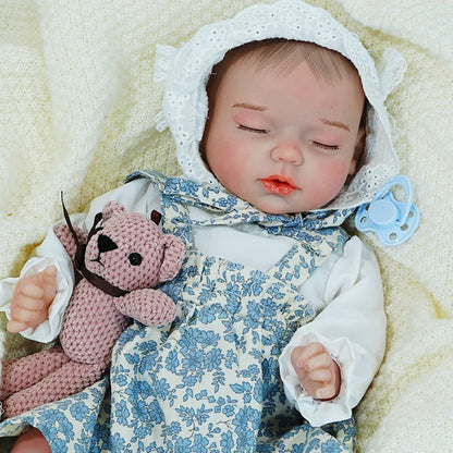 Reborn baby doll in a blue and white dress, resting on a cream blanket with eyes closed, holding a toy.