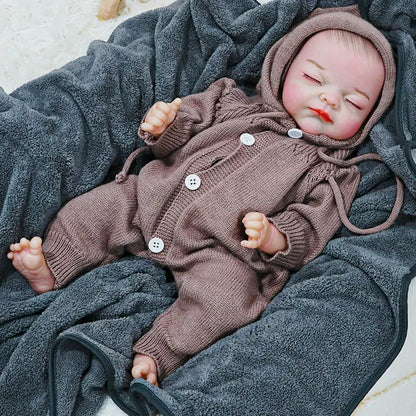 A reborn baby doll dressed in a brown hooded onesie, peacefully sleeping on a dark blanket.