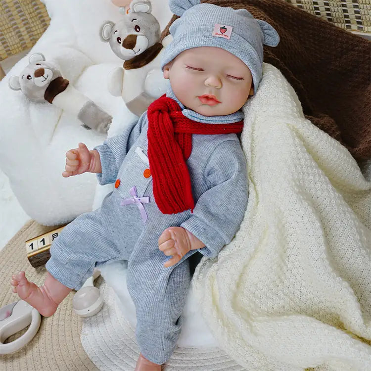 A baby doll dressed in a grey outfit with a matching hat and a red scarf, lying on a soft blanket. The doll's eyes are closed, and it is surrounded by plush bear toys, creating a cozy and peaceful scene.