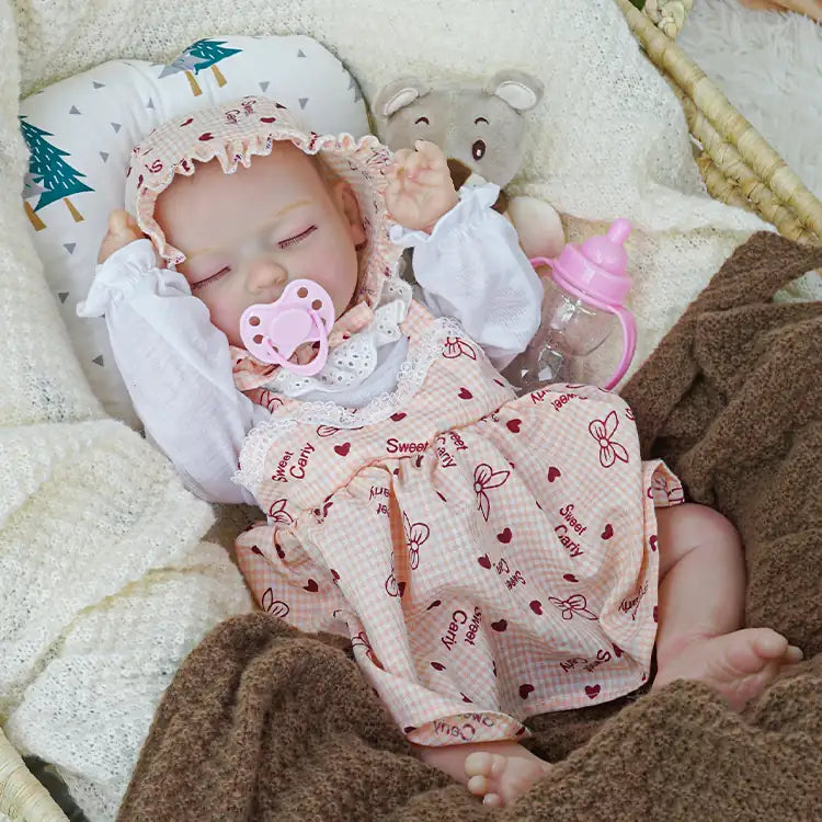 Realistic reborn doll dressed in a pink checkered outfit and white bonnet, sleeping in a wicker basket, covered with a white blanket and accompanied by a plush bear.