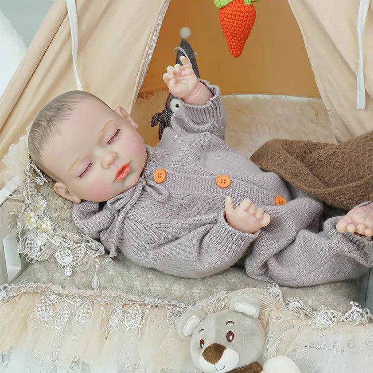 A reborn doll toy dressed in a lilac knitted outfit with a matching hood, laying on a soft white blanket, surrounded by plush toys and a tent background.