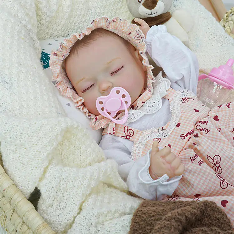Reborn doll with closed eyes, wearing a pink checkered dress and white blouse, snuggled under a brown blanket in a wicker basket, holding a pacifier, with a plush teddy bear nearby.