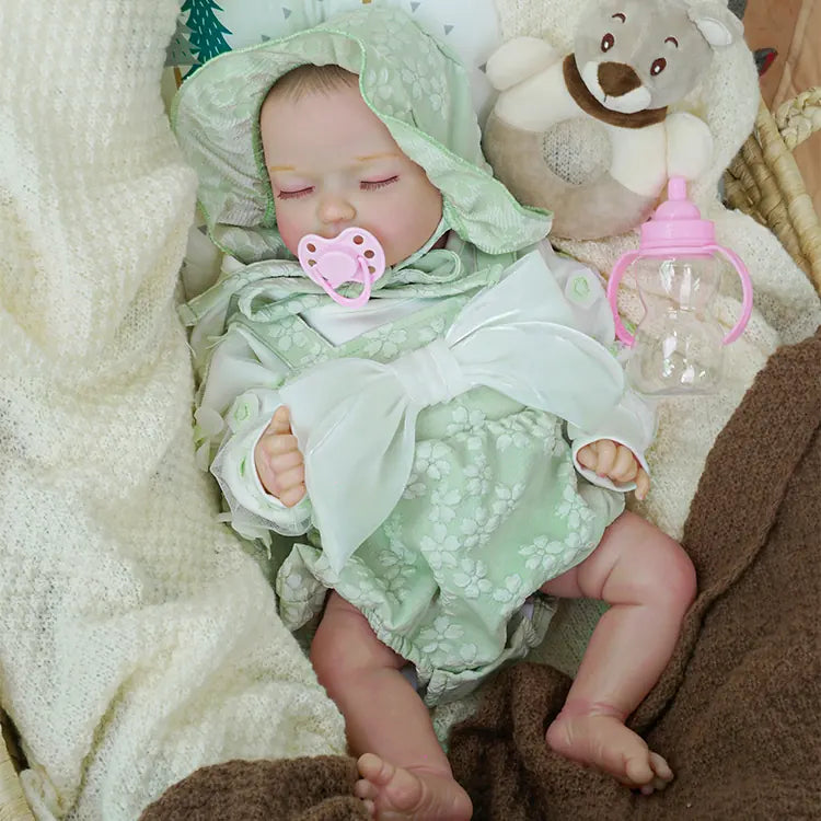 Peaceful reborn baby doll dressed in a light green floral romper and hood, resting in a wicker basket covered with a soft white blanket. Plush teddy bear and pink bottle are also in the basket, complementing the cozy setting.
